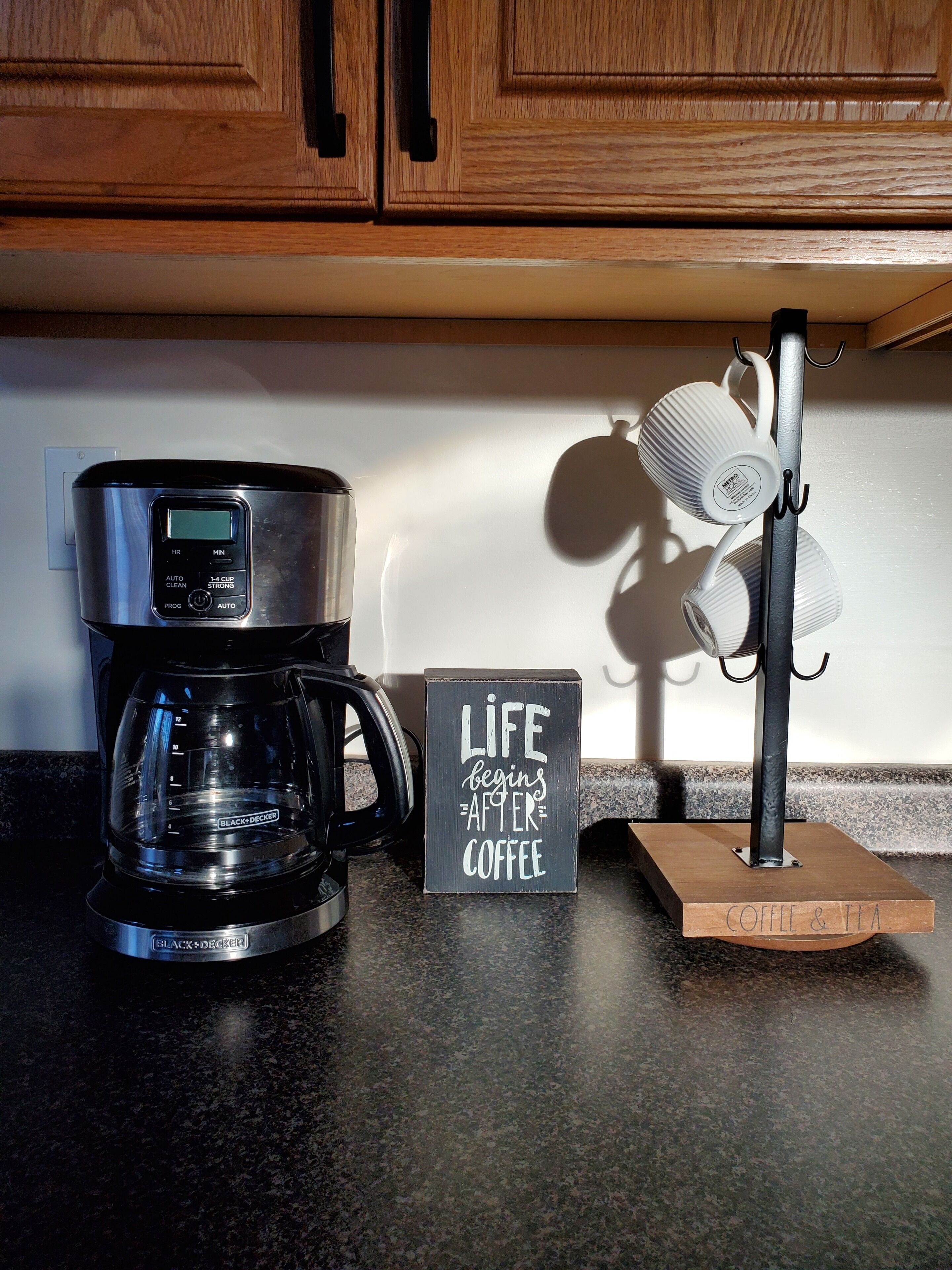 En kaffemaskine på en køkkenbordplade, et skilt med teksten 'LIFE begins after coffee', og en hylde til kopper.