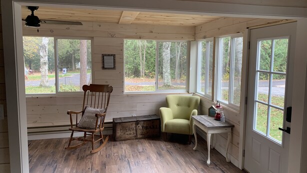 Une pièce aux murs et au plancher en bois, avec un fauteuil à bascule, un fauteuil vert, une petite table surmontée d’une lanterne rouge et de grandes fenêtres donnant sur des arbres et une route.