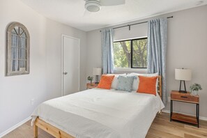 Une chambre à coucher avec un lit, une fenêtre avec des rideaux, une table de chevet avec une lampe et une porte.