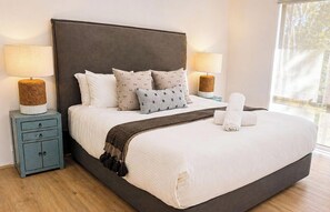 Une chambre avec un grand lit, deux tables de chevet, une commode, une fenêtre donnant sur un beau paysage et un fauteuil.