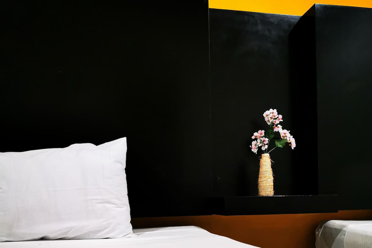 A neatly made bed with white linens, a white pillow, and a white blanket. There is a woven vase with pink flowers on a black surface.