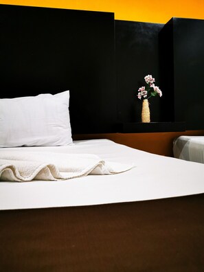 A neatly made bed with white linens, a white pillow, and a white blanket. There is a woven vase with pink flowers on a black surface.