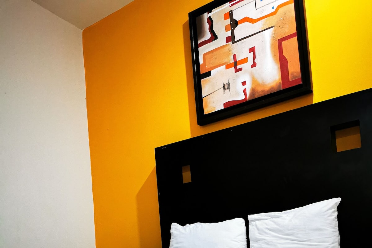 A hotel room with a bed, white pillows, a brown blanket, and a painting on the wall.