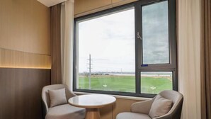 A room with two chairs and a small table, a large window with a view of a field and power lines, and a wall-mounted light fixture.