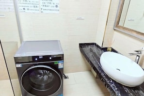 A compact hotel bathroom with a washing machine, a sink, and a mirror.