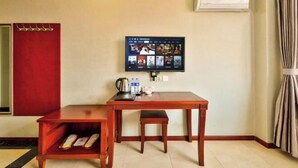A hotel room with a wooden desk, a television mounted on the wall, a red wardrobe, and an air conditioning unit.