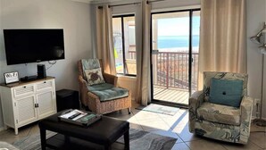 A living room with a flat-screen TV, a sofa, a chair, and a balcony with a view of the sea. - Oceanfront 3rd Floor Villa at Admiral's Row - Stunning Beach Views (Hilton Head Island)