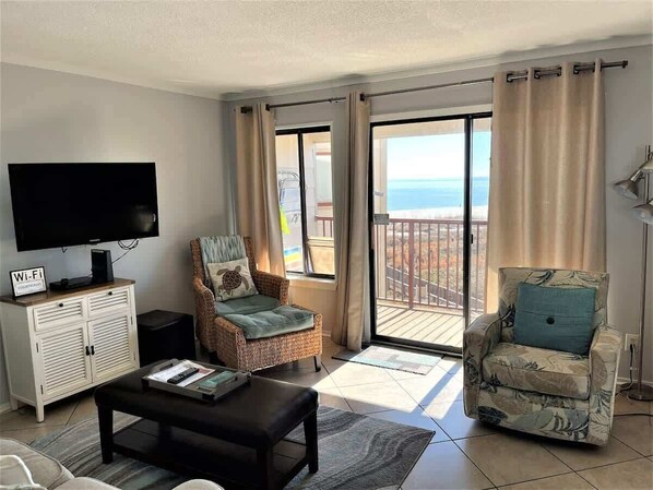A living room with a flat-screen TV, a sofa, a chair, and a balcony with a view of the sea.