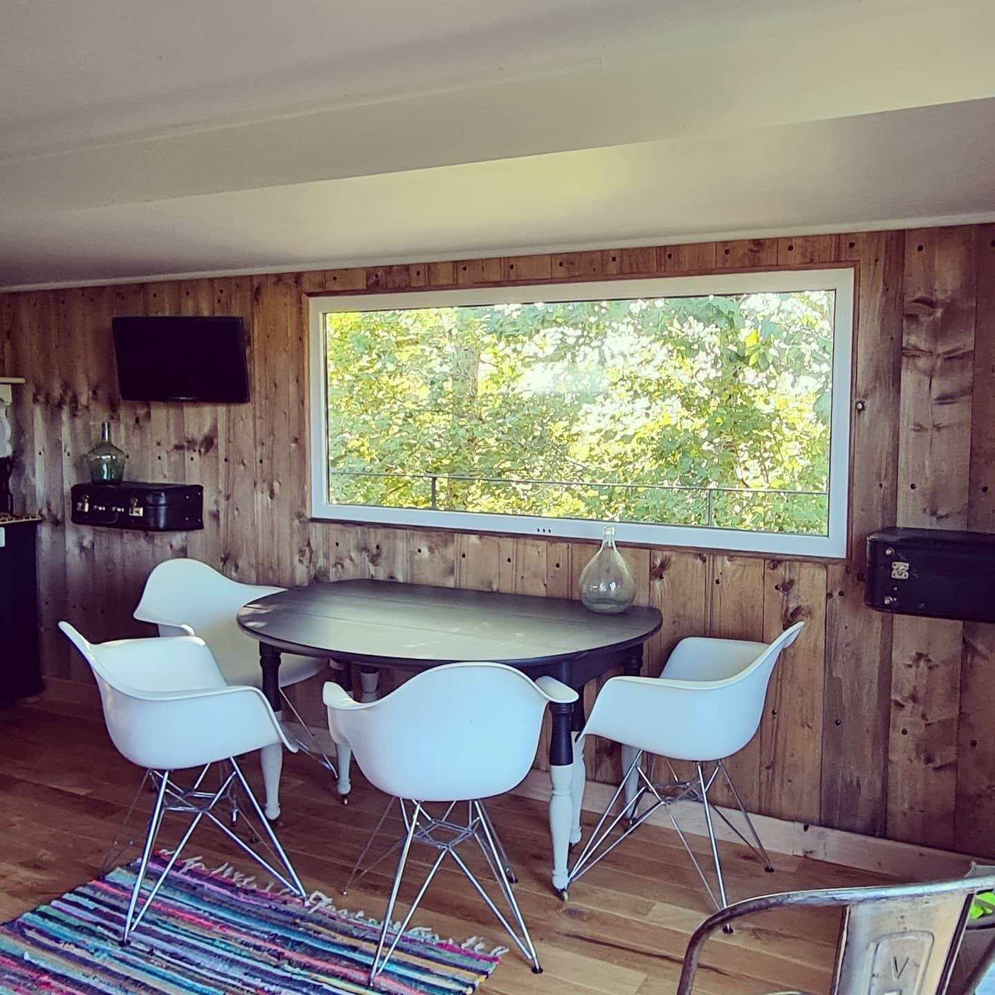 Een eetruimte met een ronde tafel, vier witte stoelen, een kleurrijk vloerkleed en een groot raam met uitzicht op bomen.