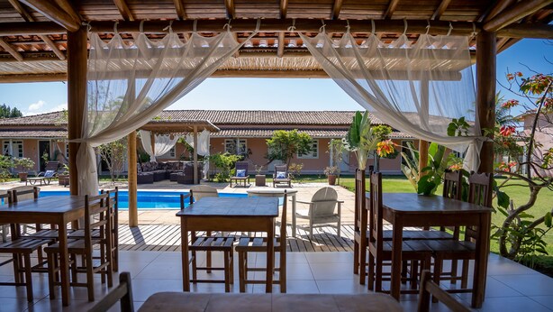 An outdoor patio with wooden tables and chairs, a pool, and a view of a house in the background. - In Trancoso, a beautiful space for up to 35 people. (Trancoso)