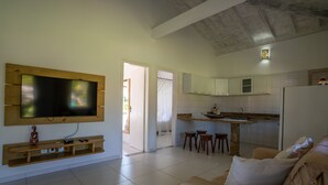 A living room with a TV, wooden entertainment unit, and a sofa. There is a kitchen area with stools and a counter. - In Trancoso, a beautiful space for up to 35 people. (Trancoso)