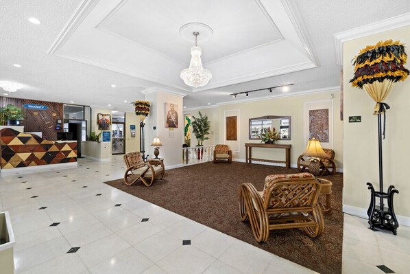A hotel lobby with a reception desk, wicker chairs, a chandelier, and a patterned carpet.