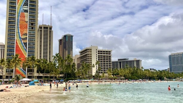 A beach with clear water, palm trees, and tall buildings in the background.