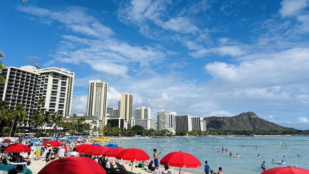 A beach with red umbrellas, people swimming, and tall buildings in the background. - Royal Kuhio 1703 - Spacious Studio w/ Stunning Mountain View in Heart of Waikiki (Waikiki)