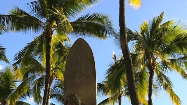 Una estatua de alguien con una tabla de surf en una playa arenosa, con palmeras y un cielo despejado.