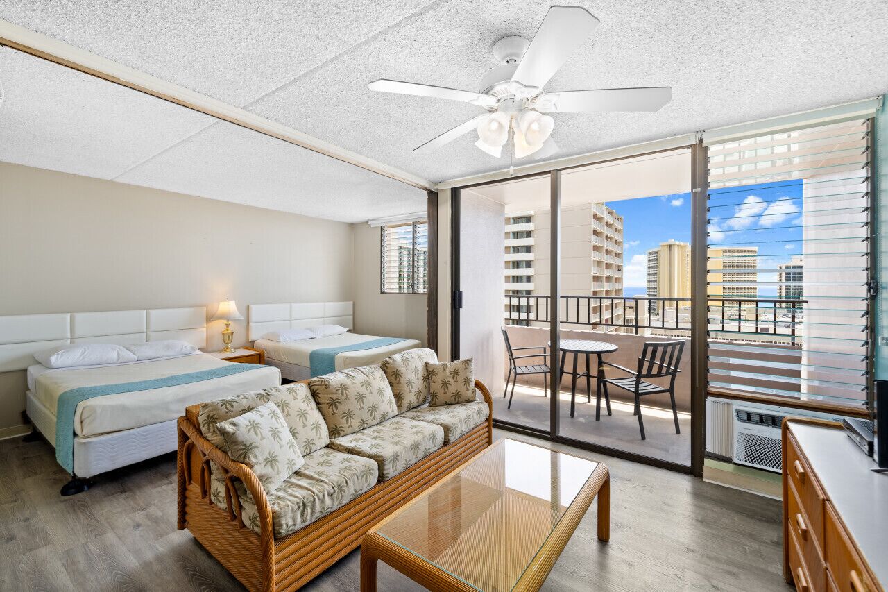 A hotel room with two beds, a sofa, a balcony with a table and chairs, and a view of the city.