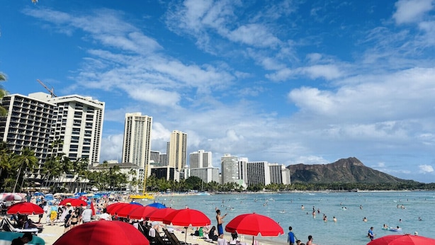 Una playa con sombrillas rojas, gente nadando y edificios altos al fondo.