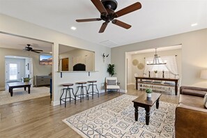 A spacious living room with a wooden floor, a patterned rug, a ceiling fan, a dining area with a chandelier, and a kitchen in the background.