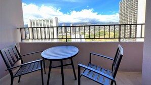 A balcony with a round table and two chairs, offering a view of tall buildings and a clear sky. - Royal Kuhio 2304 - Spacious Upgrade Studio w/ Stunning Mountain Views in Waikiki (Waikiki)