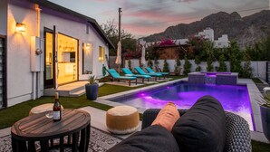 A modern outdoor pool area with a lounge chair, a table with a bottle, and a well-lit patio. - Paradise Cove with Mountain-Views Pool & Spa  (La Quinta)