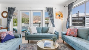 A modern living room with a teal sofa set, a round coffee table, and a large window with curtains. - Paradise Cove with Mountain-Views Pool & Spa  (La Quinta)