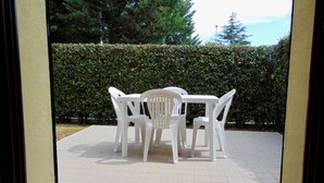 Eine Terrasse mit Tisch und Stühlen, einem gefliesten Boden und einem Hecken im Hintergrund.