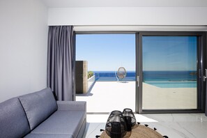 A modern living room with a grey sofa, a large sliding glass door leading to a balcony with a view of the sea and a pool. - Brand new villa in Falasarna★Seaview★Infinity pool (Phalasarna)