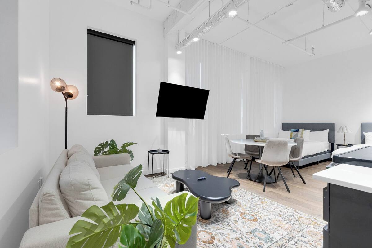A modern, well-lit hotel room with a large bed, a sofa, a dining area, and a TV mounted on the wall.