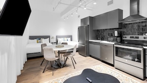 A modern kitchen with stainless steel appliances, a dining area with a round table and chairs, and a separate bedroom with a bed and nightstand.