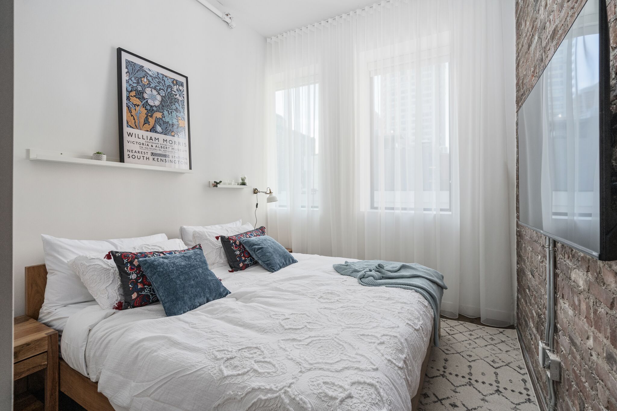 A bedroom with a bed, pillows, a bedside table, a wall-mounted TV, and a window with sheer curtains.