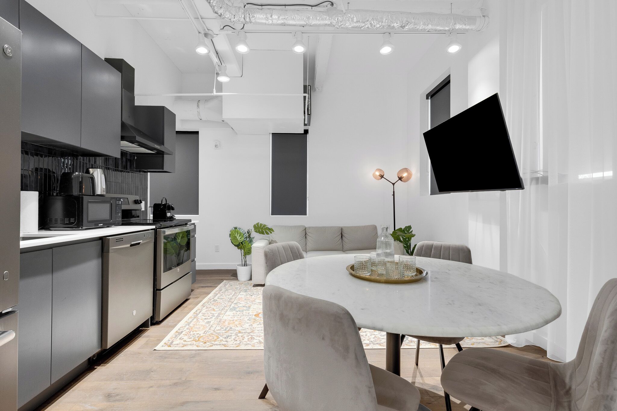 A modern kitchen with stainless steel appliances, a dining area with a round table and chairs, and a flat-screen TV mounted on the wall.