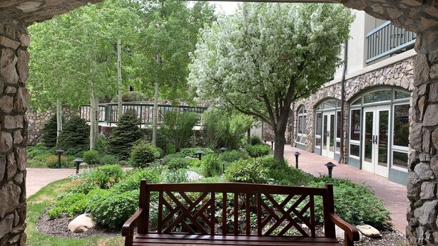 A stone archway leading to a garden with a wooden bench, surrounded by lush greenery and a stone building with arched windows. - Beaver Creek Ski-in/Ski-out Condo (Avon)