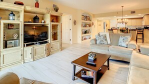 A living room with a sofa, coffee table, and TV stand. There is a kitchen in the background.