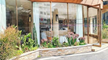 A hotel entrance with a glass facade, a stone planter with plants, and a clear view of the street outside.