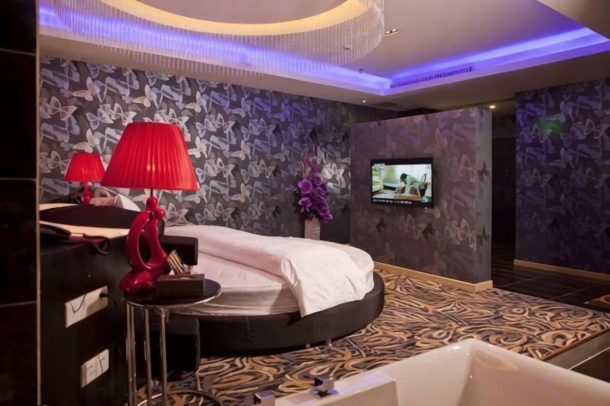 A modern hotel room with a curved bed, a TV, a red lamp, and a patterned carpet.