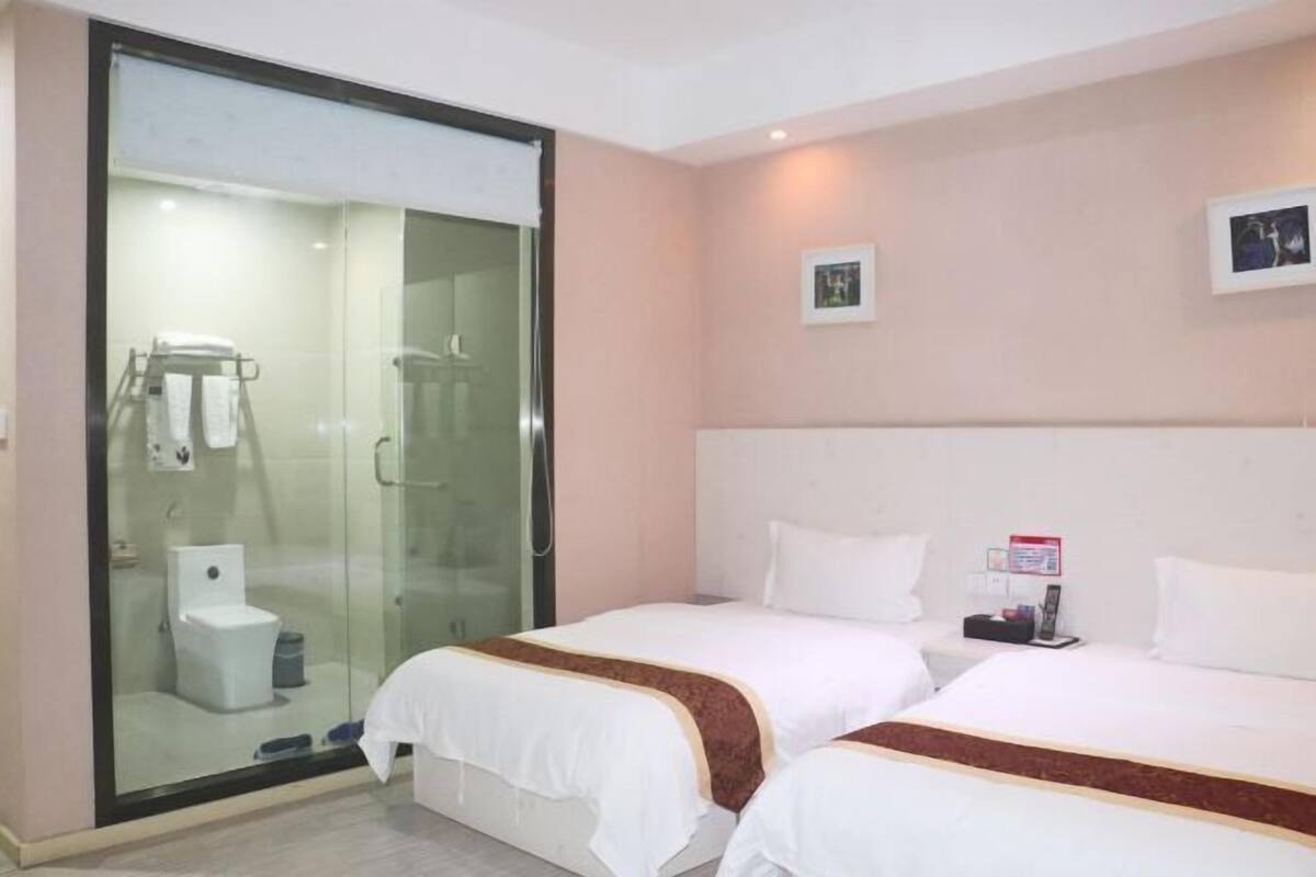 A hotel room with two beds, a glass shower enclosure, and a small bedside table with a phone and a book.