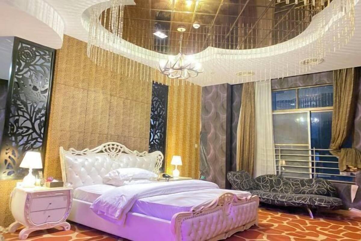 A bedroom with a large bed, a nightstand, a sofa, a chandelier, and a window with curtains.