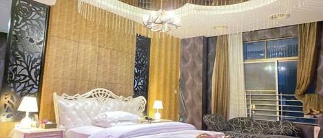 A bedroom with a large bed, a nightstand, a sofa, a chandelier, and a window with curtains.
