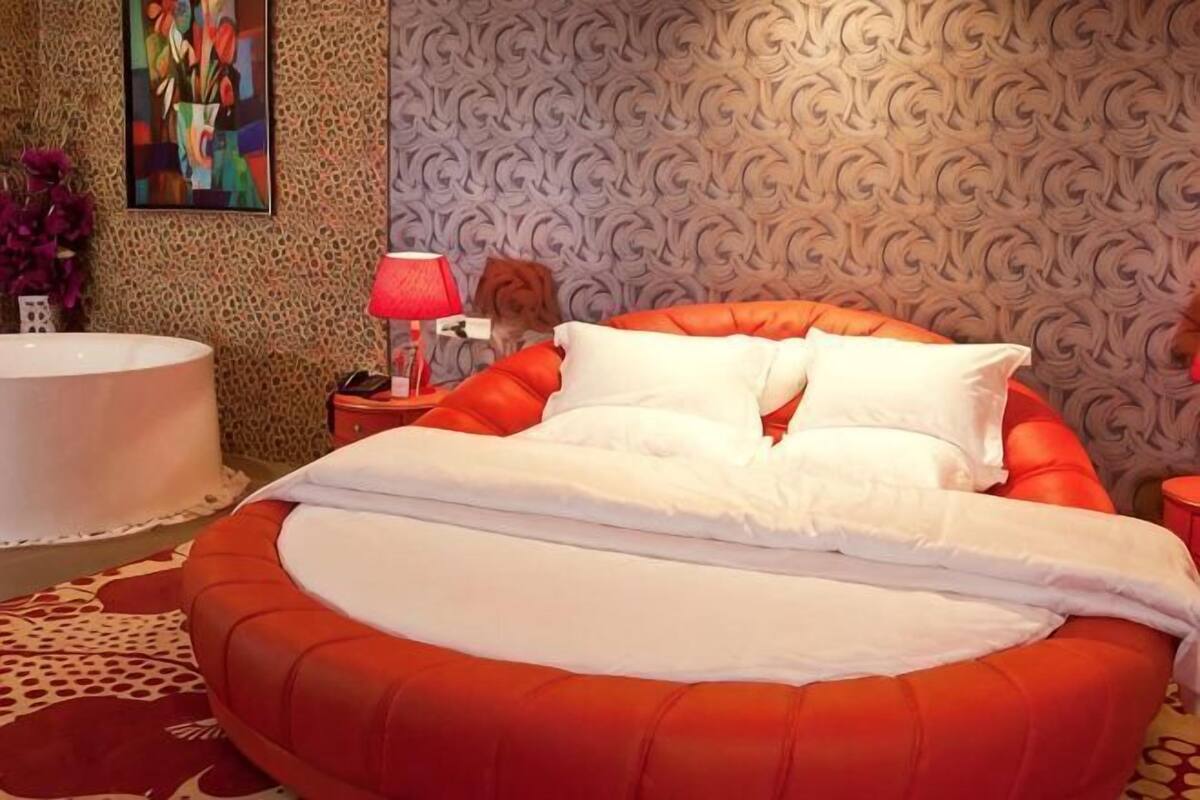 A bedroom with a large bed, a round red bed frame, a white bathtub, and a patterned wallpaper.