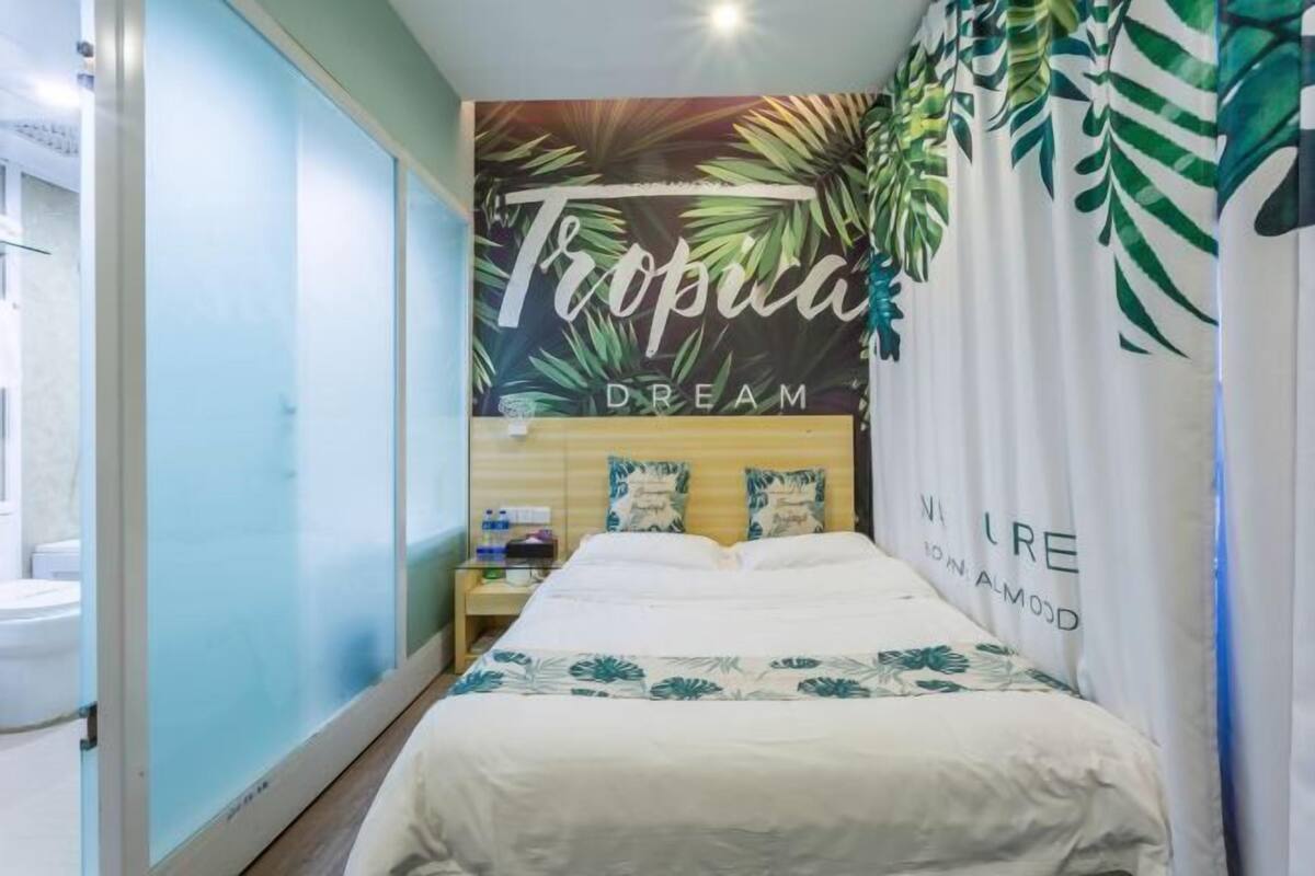 A bedroom with a bed, a nightstand, a wall with a tropical-themed mural, and a curtain with a leaf pattern.