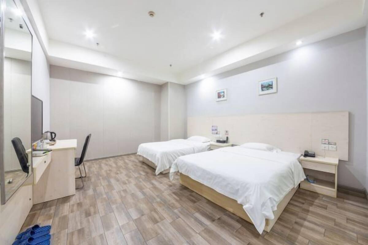 A hotel room with two single beds, a desk with a chair, a small bedside table with a lamp, and a mirror.