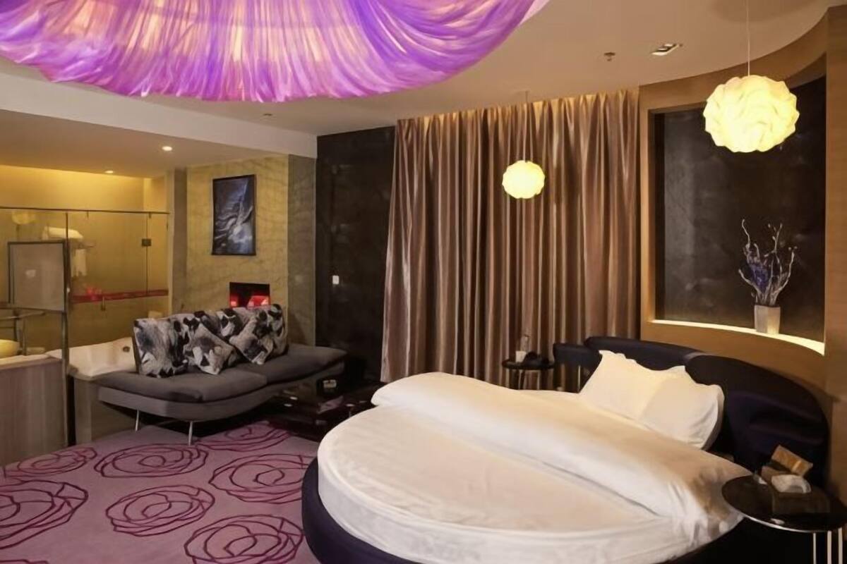 A modern hotel room with a large bed, a sofa, a glass-enclosed shower, and a view of the city.