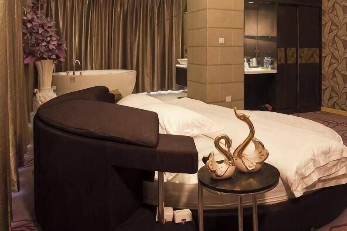 A hotel room with a bed, a round black chair, a small round table with a decorative item, a bathtub, and a mirror.