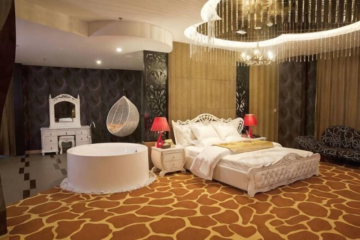 A bedroom with a circular bathtub, a bed with white bedding, a hanging chair, and a chandelier.