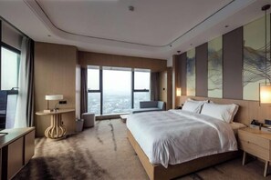 A modern hotel room with a large bed, a desk, a chair, and a view of the city through large windows.