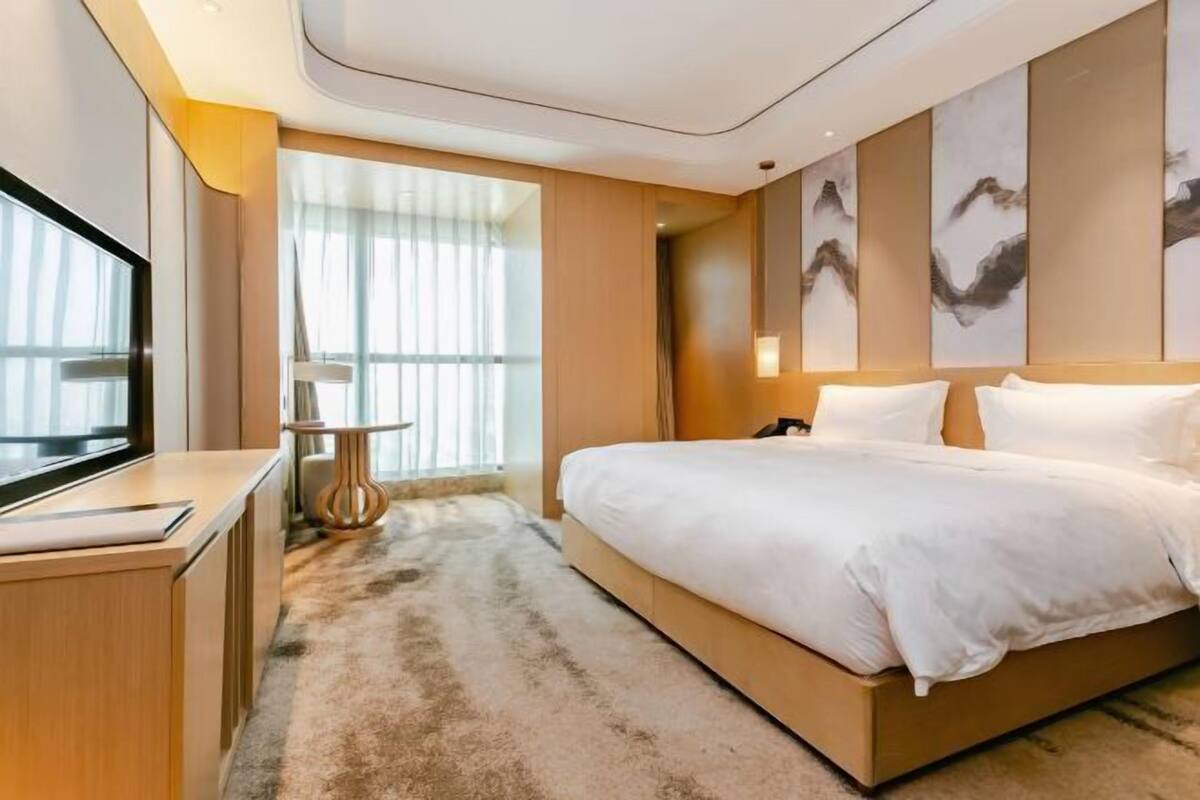 A modern hotel room with a large bed, a TV, a bedside table, and a window with sheer curtains.