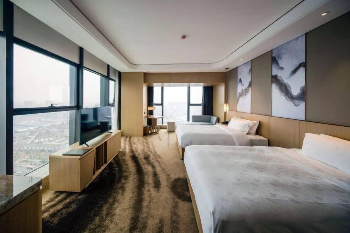 A modern hotel room with large windows, a flat-screen TV, two beds, a sofa, and artwork on the walls.