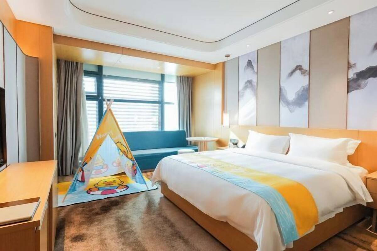 A hotel room with a large bed, a small desk, a chair, a window with curtains, and a wall with abstract art.