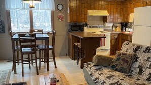 Un salon chaleureux avec une table à manger en bois, un canapé agrémenté de coussins à motifs animaliers et un coin cuisine doté de meubles en bois et d’un réfrigérateur.