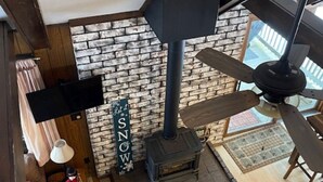 Une pièce avec une cheminée en briques, un ventilateur de plafond et une table en bois sur laquelle se trouve une bouteille.